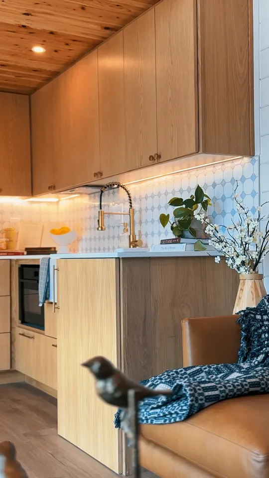 Modern kitchen with wooden cabinets and ceiling, white countertop, and white tile backsplash with geometric patterns, featuring a gold faucet, plants, books, and a vase with white flowers next to a brown leather chair with a blue patterned blanket.