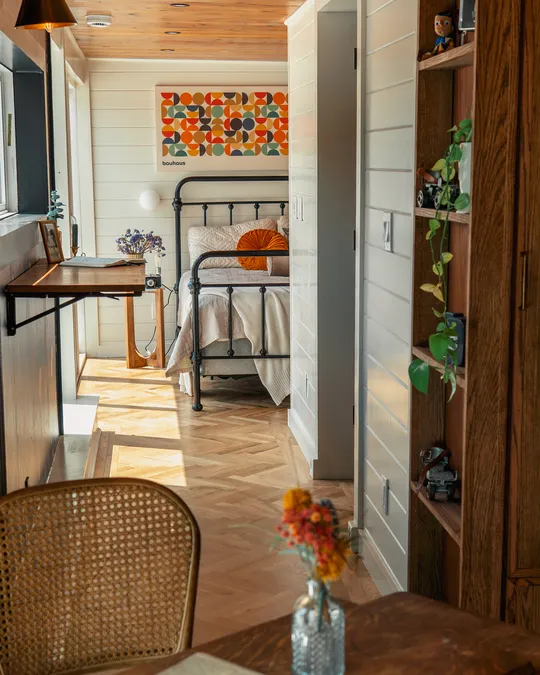 A cozy bedroom with a black metal bed frame, white bedding, and an orange round cushion, next to a white wall with a colorful geometric poster labeled 'bauhaus'. A small wooden side table holds a vintage camera and flowers. The room has light wood parquet flooring, a wooden shelf with plants and toys, and a window desk with a lamp. A rattan chair and a wooden table with a small vase of flowers are visible in the foreground.
