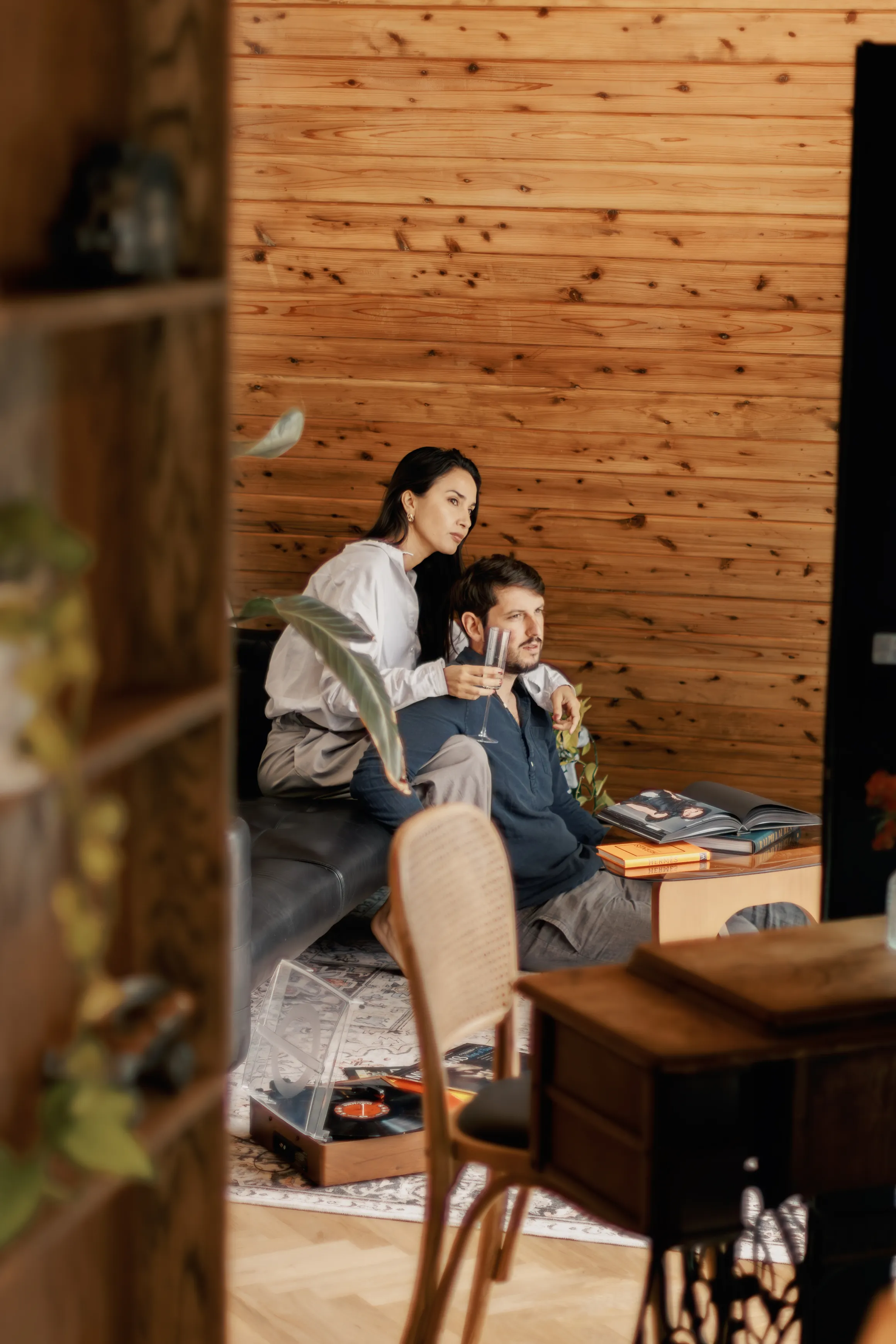 A couple sitting in a cozy wooden room with the woman holding a glass and leaning on the man's shoulder as they watch something together; a record player and books are visible nearby.