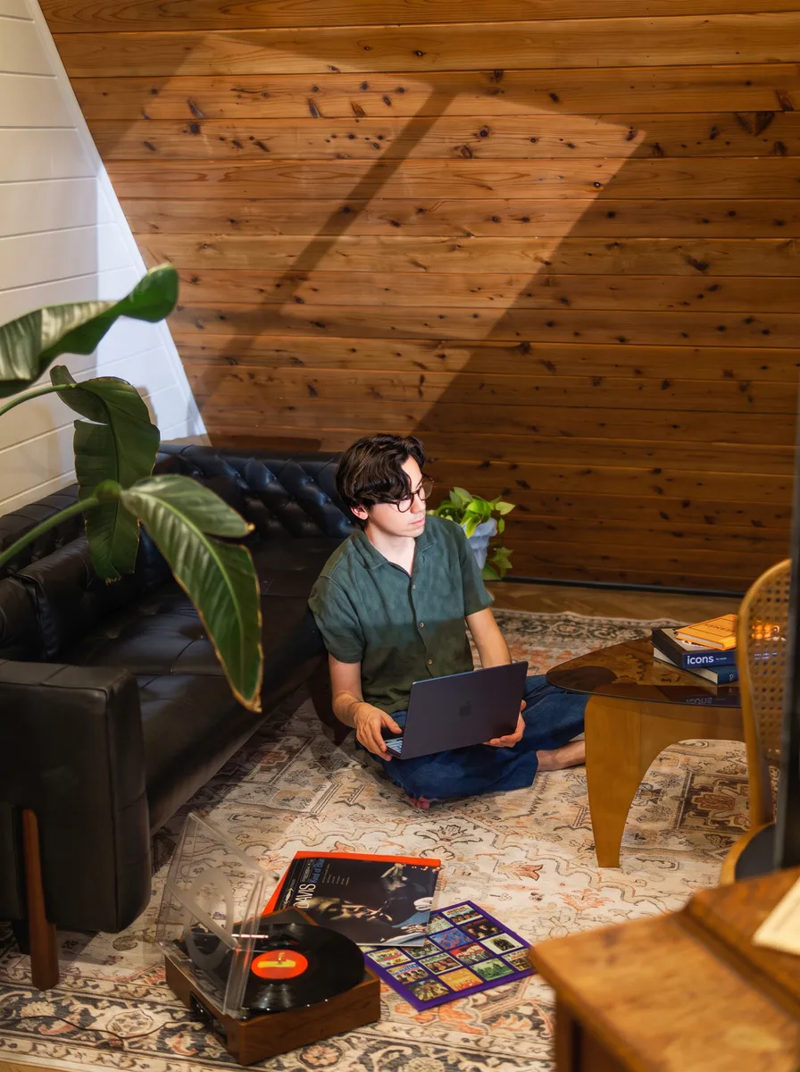 Young person with glasses sitting cross-legged on a patterned rug in a cozy room with wood-paneled walls, using a laptop, near a black leather couch, a coffee table with books, a record player with vinyl records, and a large green plant.