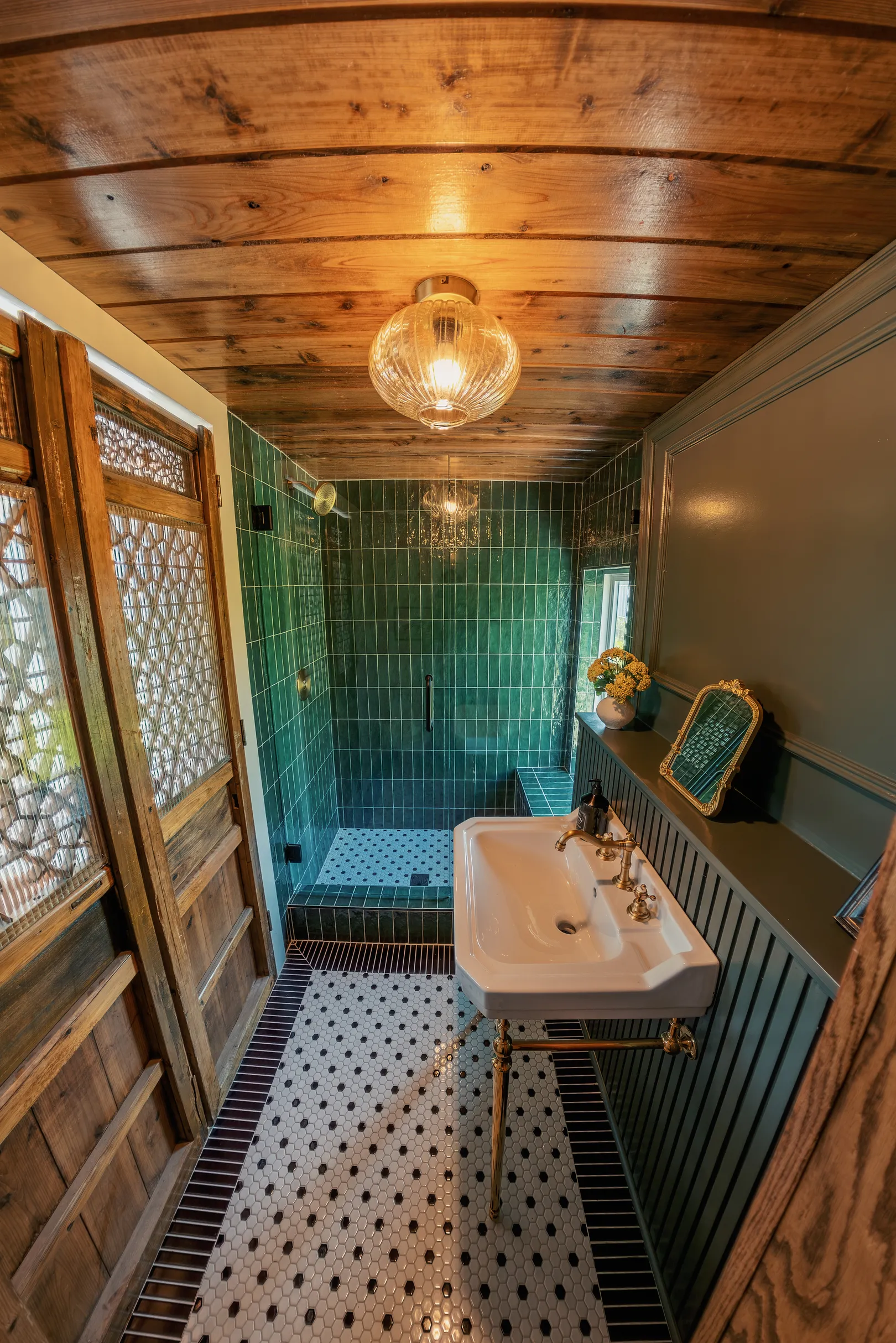 Small bathroom with wooden ceiling and double wooden doors, featuring a white sink with brass fixtures on gold legs, green tiled shower with built-in bench, and black-and-white hexagonal floor tiles bordered by dark rectangular tiles. A small ornate mirror and vase with flowers are on a green paneled ledge beside the sink.