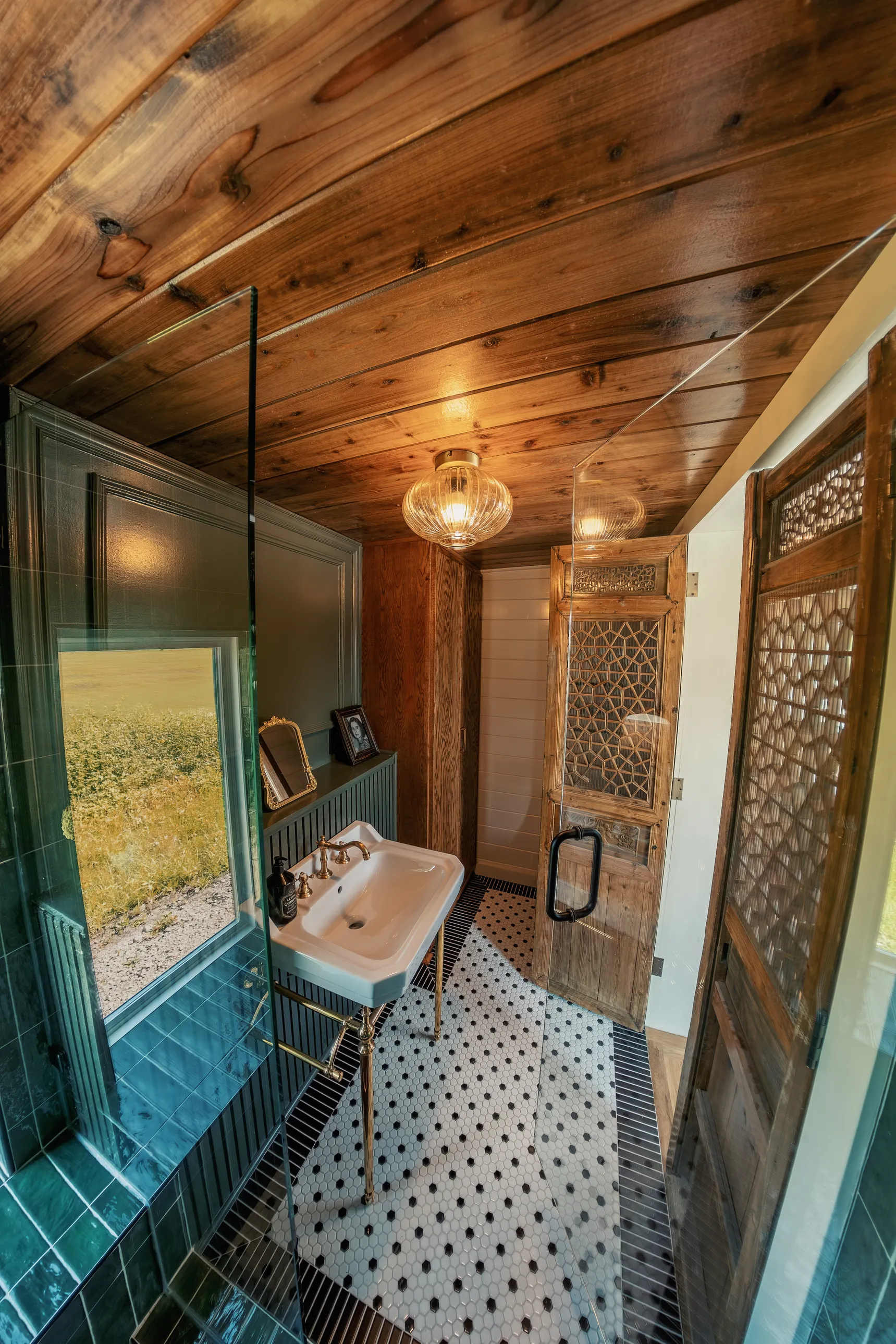 Small bathroom with hexagonal tiled floor in black and white, wooden ceiling, white sink with brass fixtures, a window showing an outdoor field, and carved wooden doors, illuminated by a round ceiling light.