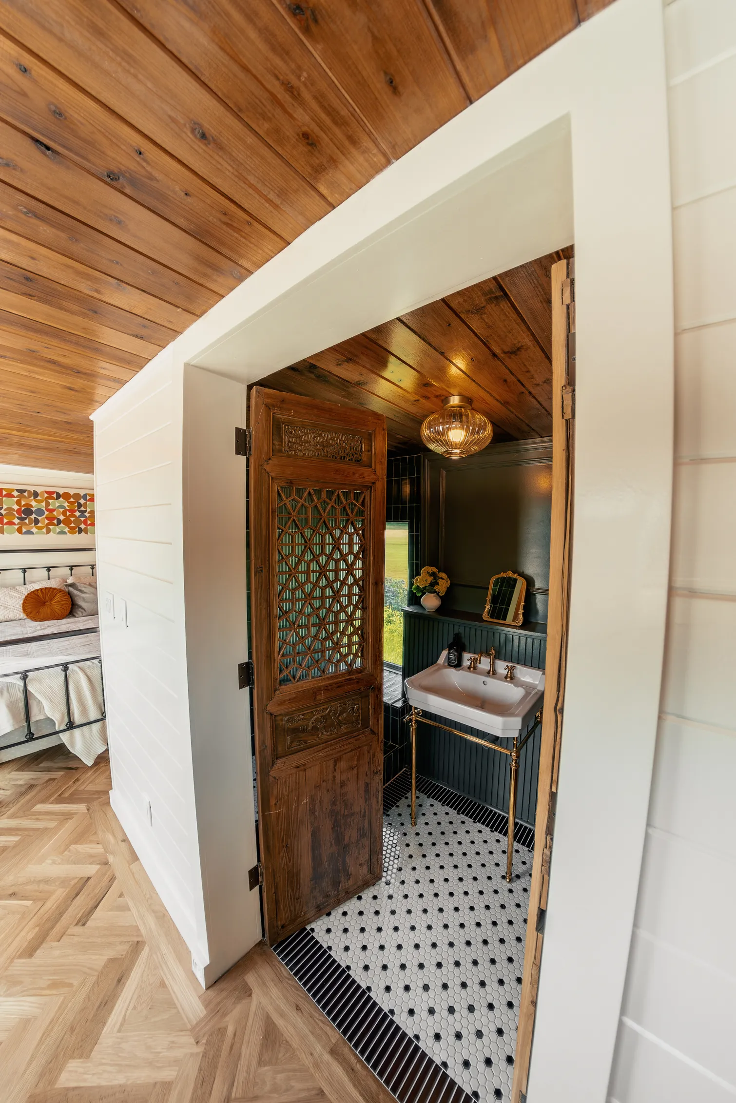 Interior view of a cozy bedroom with a visible black metal bed frame, white bedding, and a colorful retro wall art piece, adjacent to a bathroom with a wooden carved door, white sink with gold fixtures, small mirror, flowers in a vase, dark green wainscoting, and a tiled black-and-white floor.