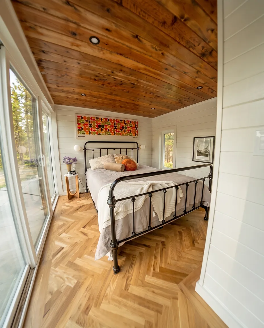 Cozy bedroom with a black metal bed frame, light bedding, and decorative pillows, wooden herringbone floor, wooden ceiling, white shiplap walls, a colorful abstract artwork above the bed, and large sliding glass doors letting in natural light.