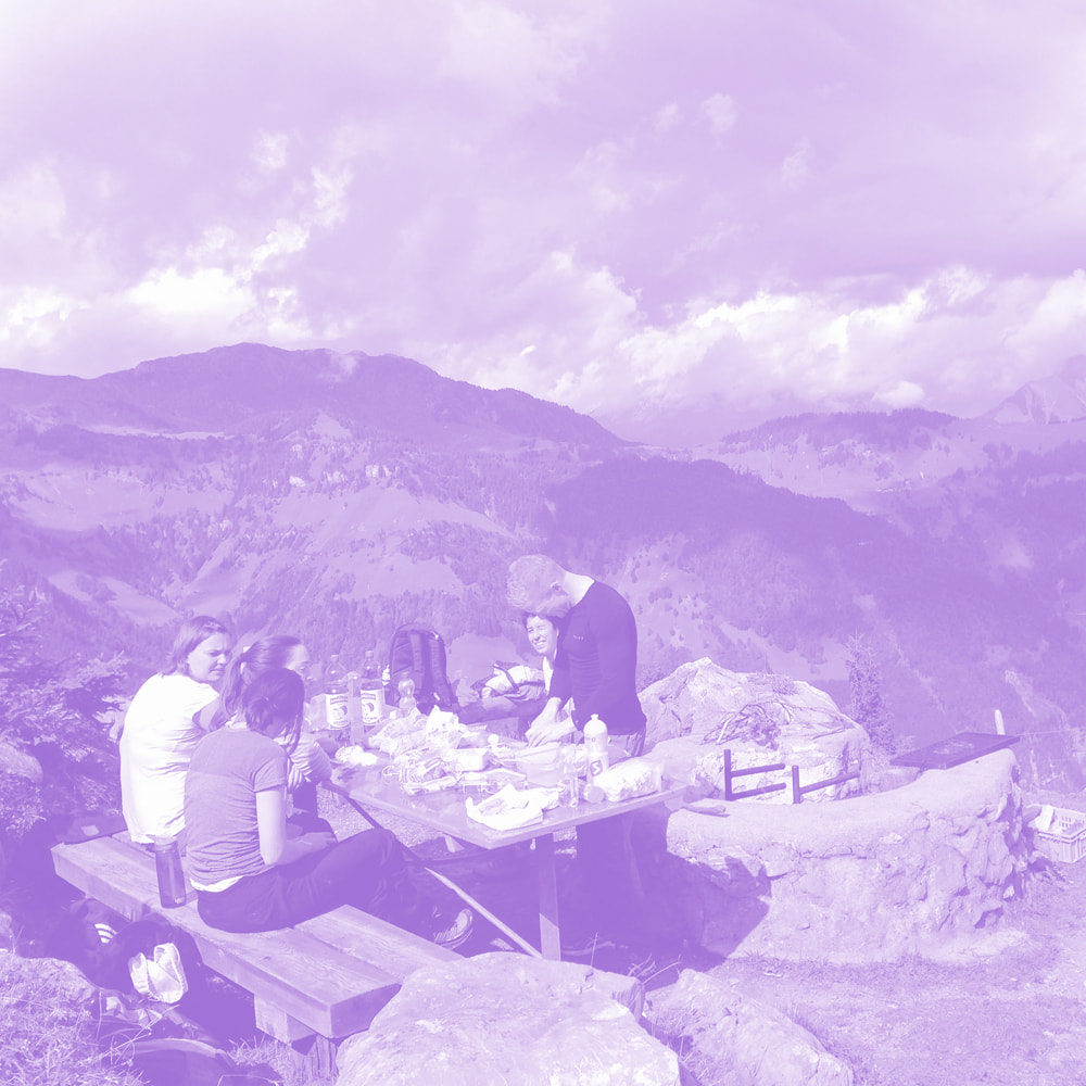 Group of people having a picnic at a wooden table with food and drinks, set against a mountainous landscape under a partly cloudy sky.