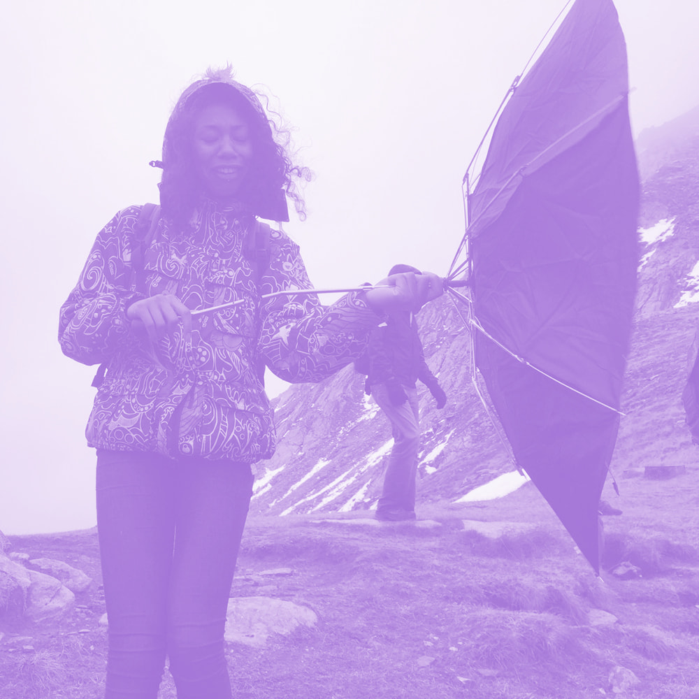 Person outdoors holding an inverted umbrella on a windy mountain trail with rocky terrain and patches of snow.