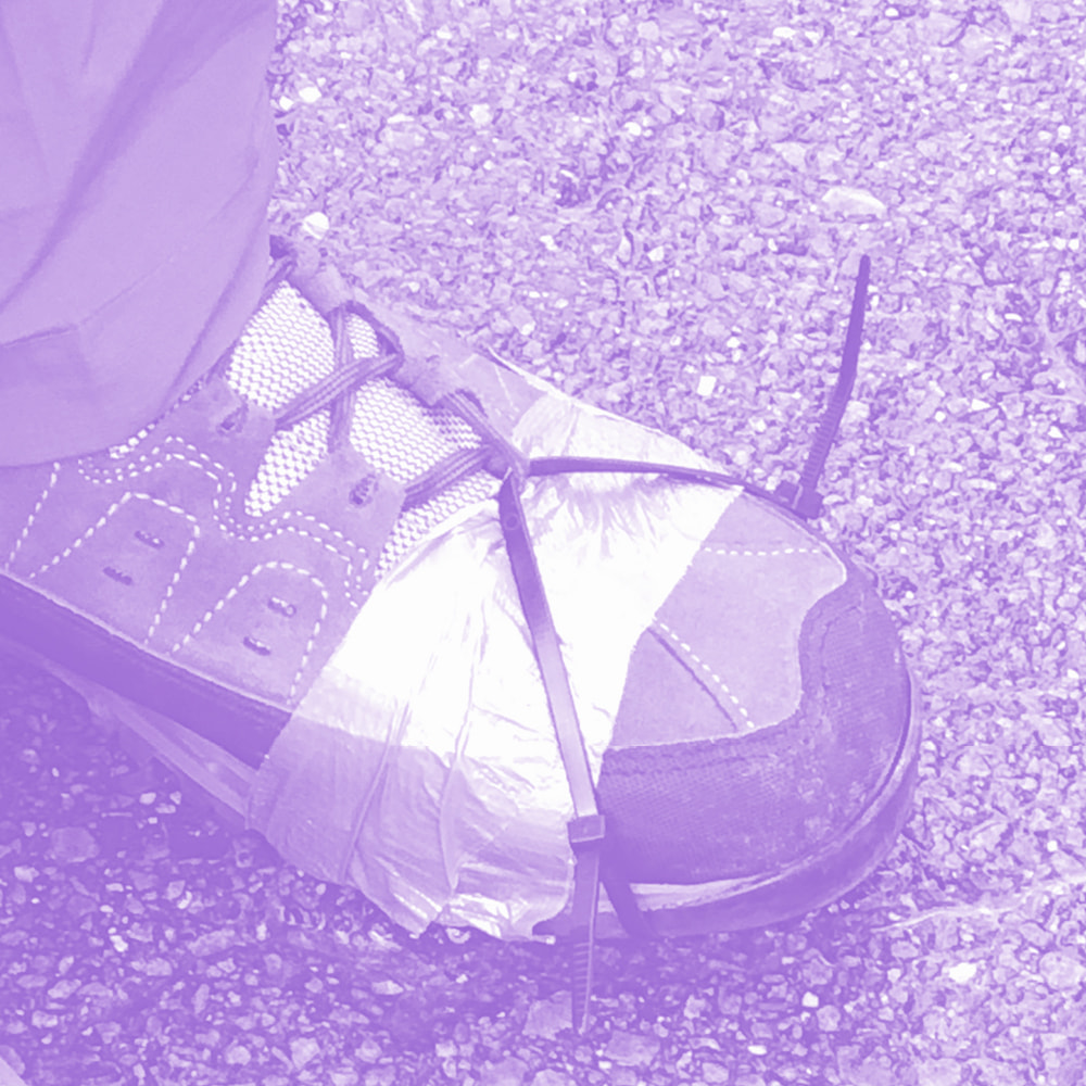 Close-up of a hiking boot wrapped with silver duct tape and secured with black zip ties on gravel ground.