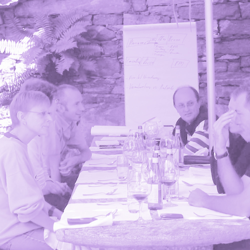 Group of people sitting at a long table with bottles and glasses, having a discussion in a room with stone walls and a flipchart.