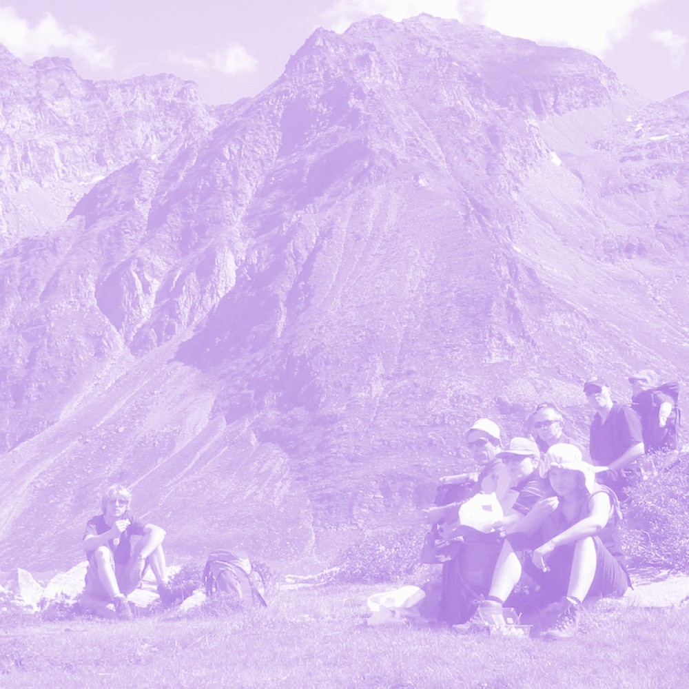 Group of hikers resting on grass with backpacks in front of a large mountain.