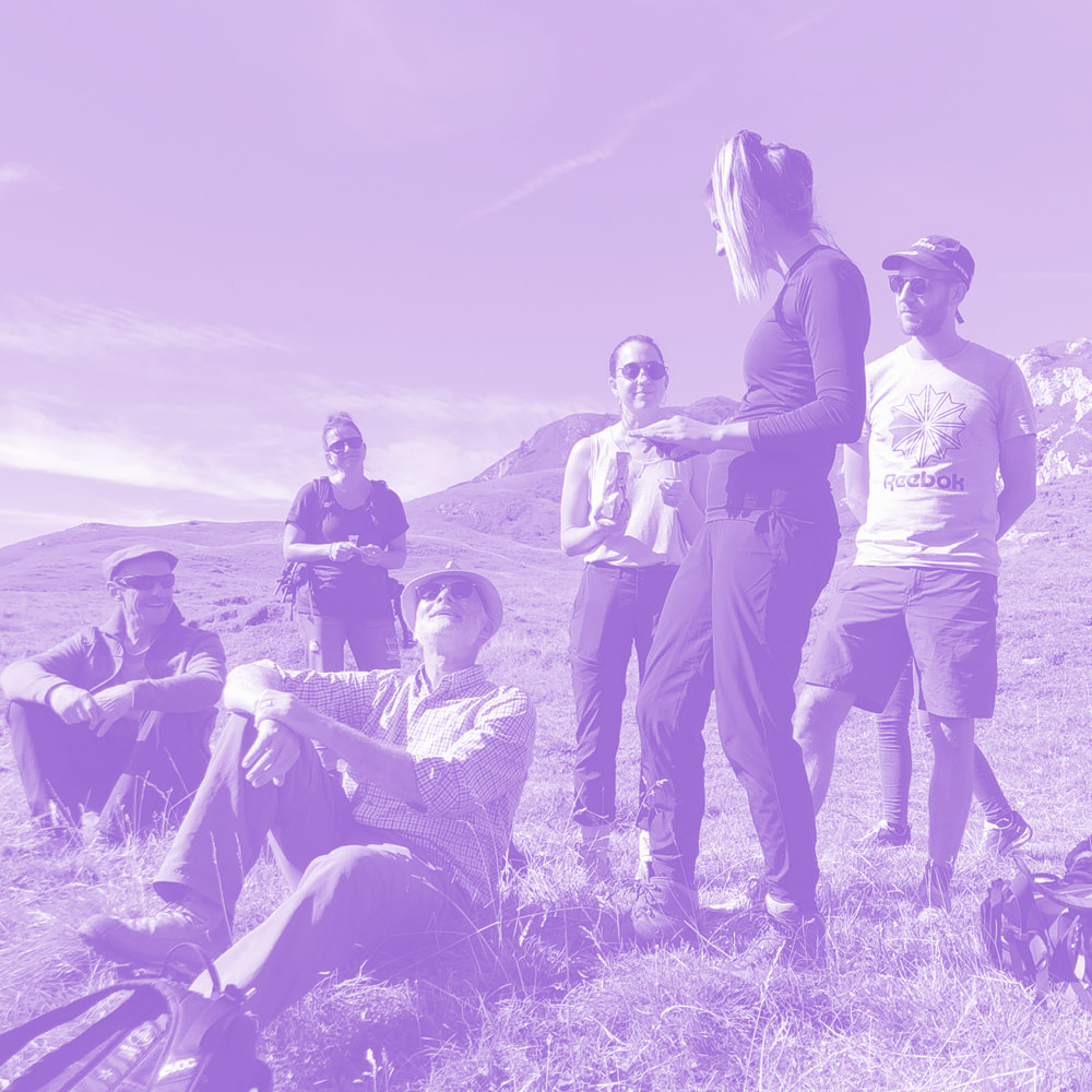 Group of six hikers resting and chatting on a grassy hillside with mountains in the background.