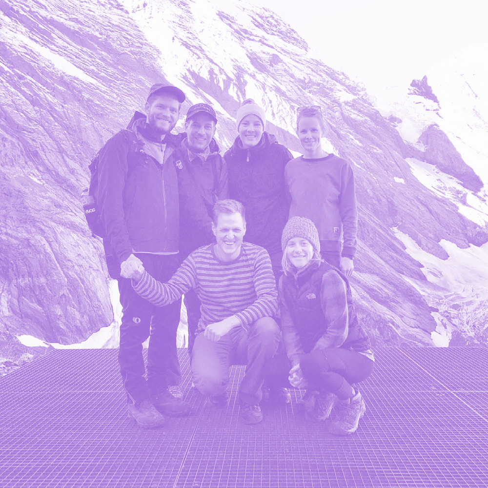 Group of six hikers smiling on a metal platform with rocky snow-covered mountain cliffs in the background.