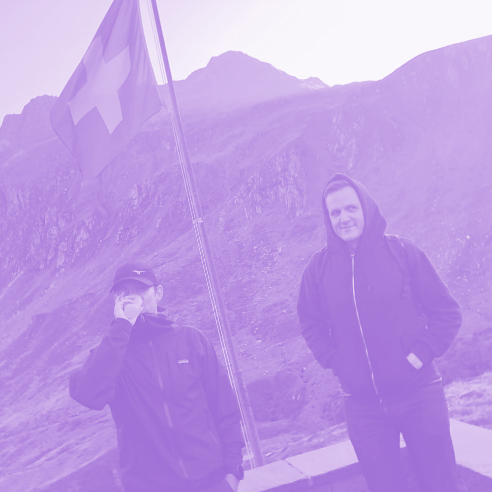 Two men, one drinking from a cup, standing near a Swiss flag on a mountainous background.