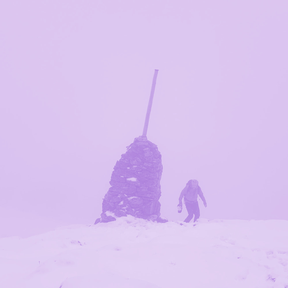 Person hiking near a stone cairn with a vertical pole on a snowy mountain in foggy weather.