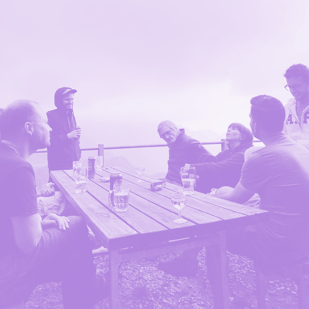Group of six people sitting and standing around a wooden picnic table outdoors with drinks, enjoying a conversation against a mountainous backdrop.