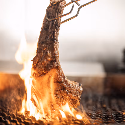 Close-up of a grilled ribeye steak being held over flames on a barbecue grill.