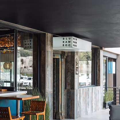 Outdoor seating area of a modern cafe with wooden chairs, potted plants, and a white sign reading 'BLU BAM BOO'.