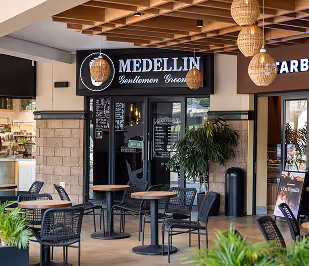 Outdoor seating area with tables and chairs in front of Medellin Gentlemen Grooming shop with wooden ceiling and hanging lamps.