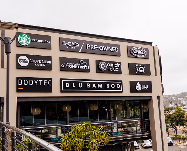 Exterior building wall with multiple business signs including Starbucks, Cars Bassonia Pre-Owned, The Crazy Store, Crisp & Clean Laundry, Comaro View Optometrists, Curtain Club, Absolute Pets, Bodytec, Blu Bamboo, and V Bar.