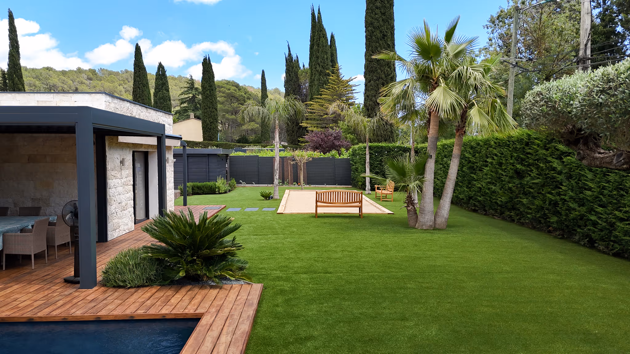Modern backyard with a wooden deck, outdoor dining area, green lawn, palm trees, and wooden benches around a sandy bocce court.