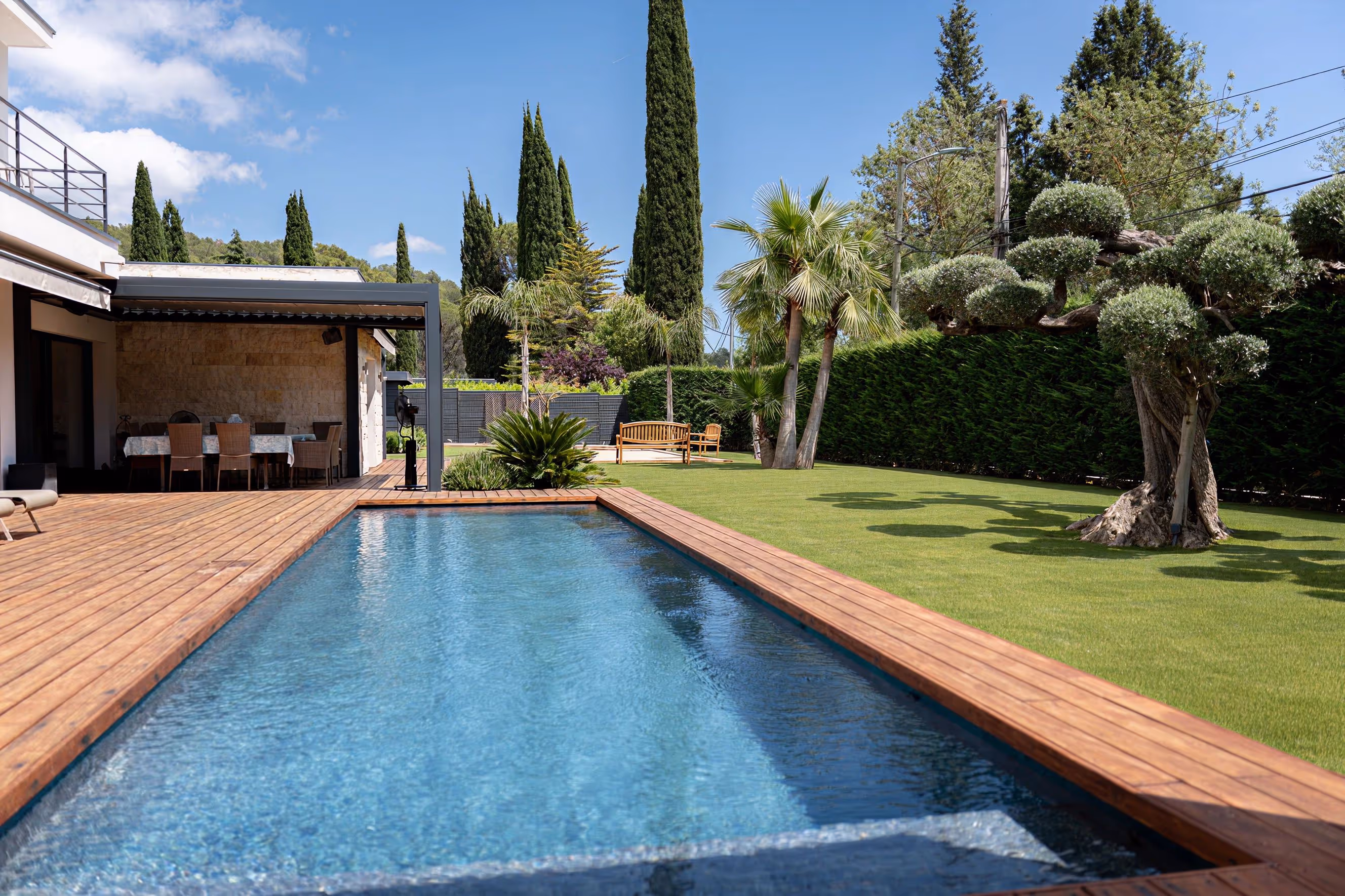 Modern backyard with a rectangular swimming pool, wooden deck, lawn, palm trees, and outdoor dining area under a pergola.