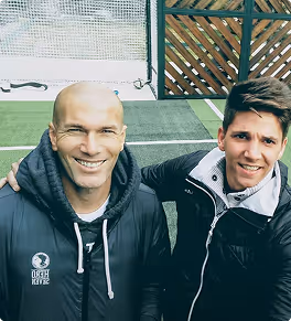Two men smiling and posing together on a sports field with netting and wooden fencing in the background.