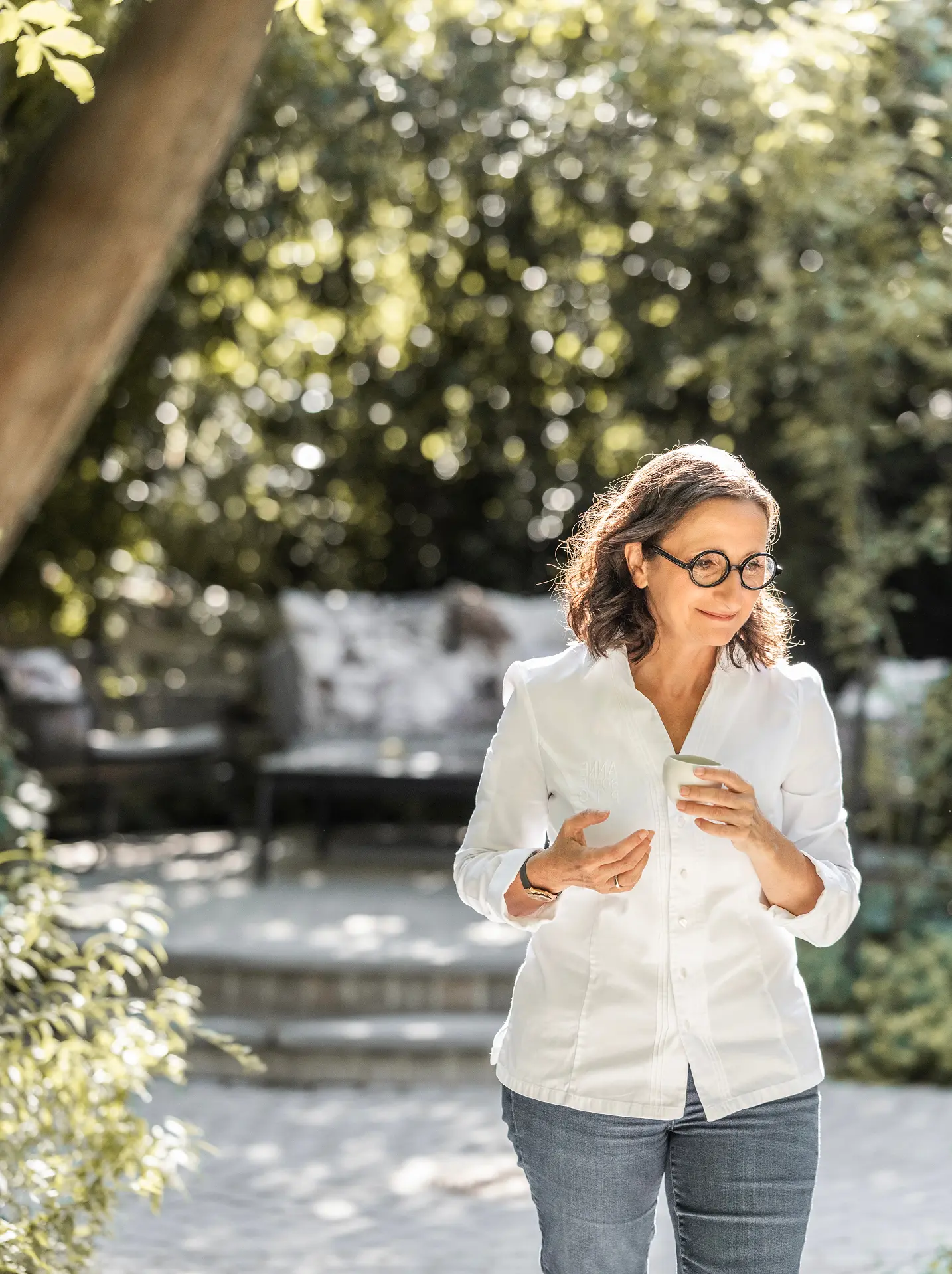 Femme portant une veste blanche et des lunettes rondes, tenant une tasse dans un jardin ensoleillé.