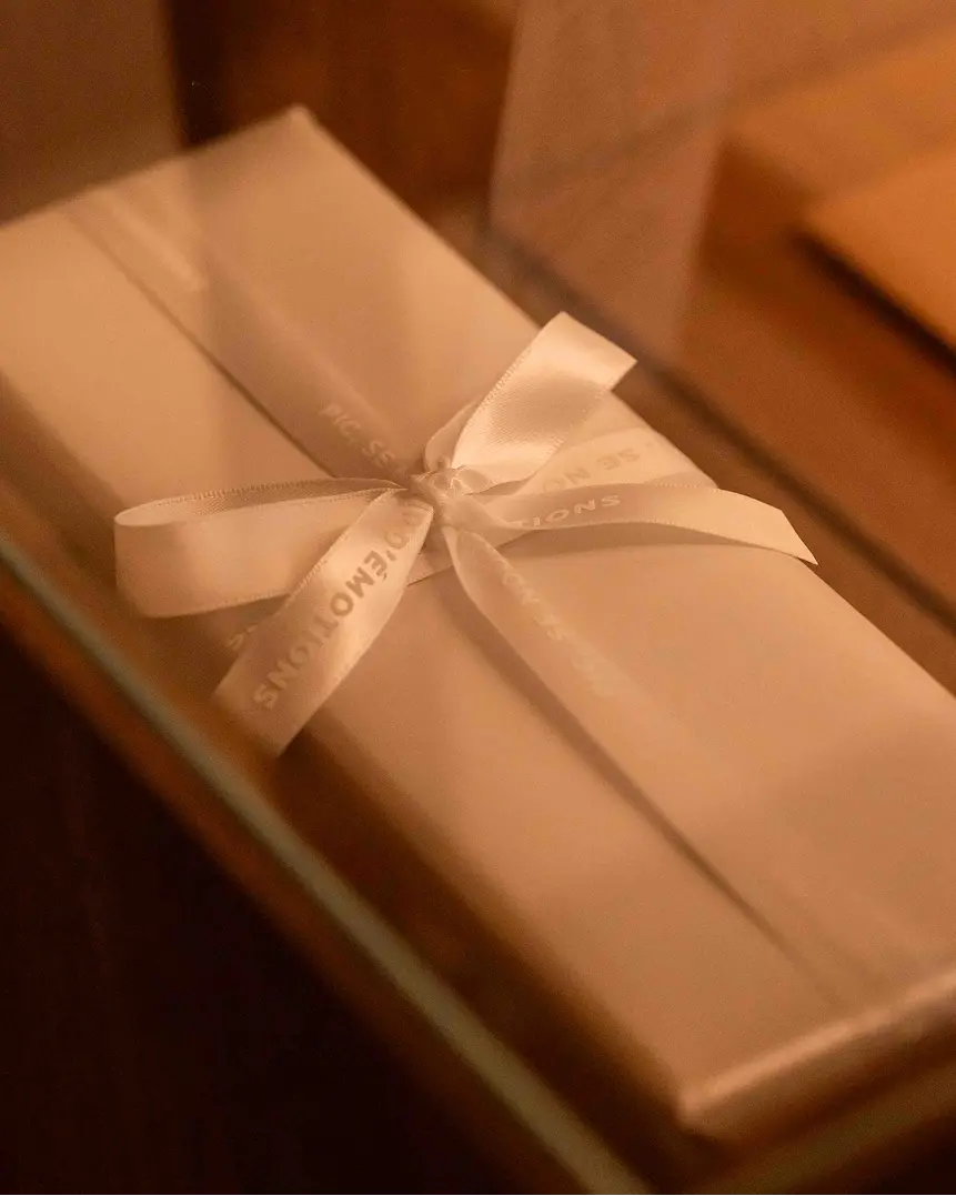Rectangular beige gift box with a white ribbon tied in a bow.