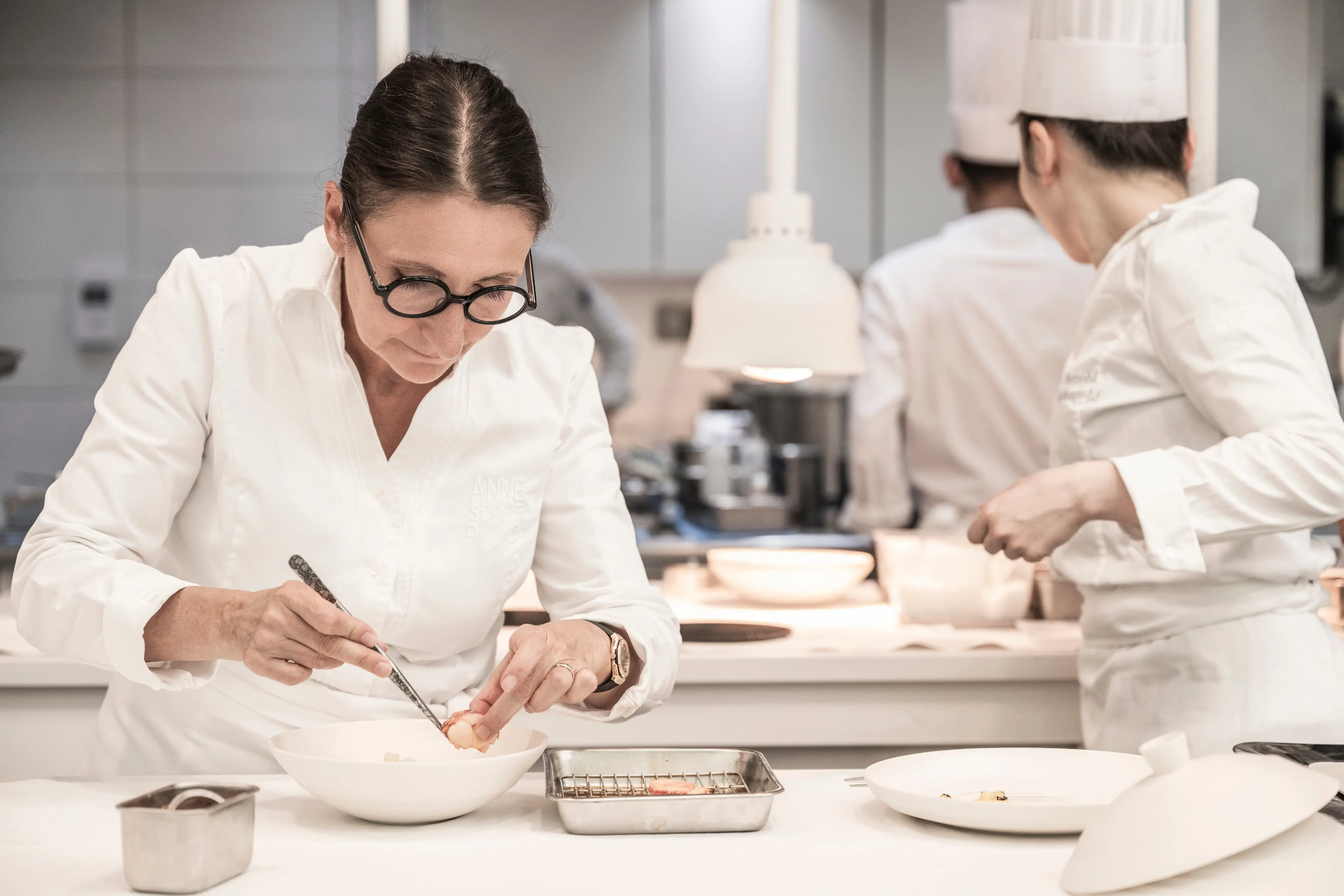 Chef en blouse blanche et lunettes, concentrée à décorer un plat dans une cuisine professionnelle.