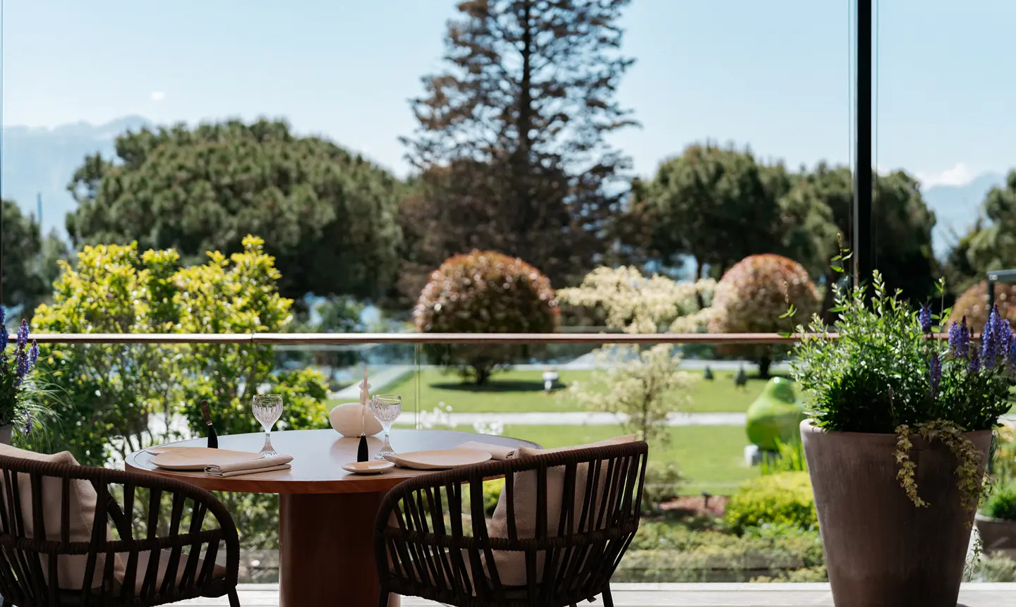 Terrasse mit einem Holztisch für zwei Personen, umgeben von Korbstühlen, mit Blick auf einen grünen Garten und Bäume im Hintergrund.