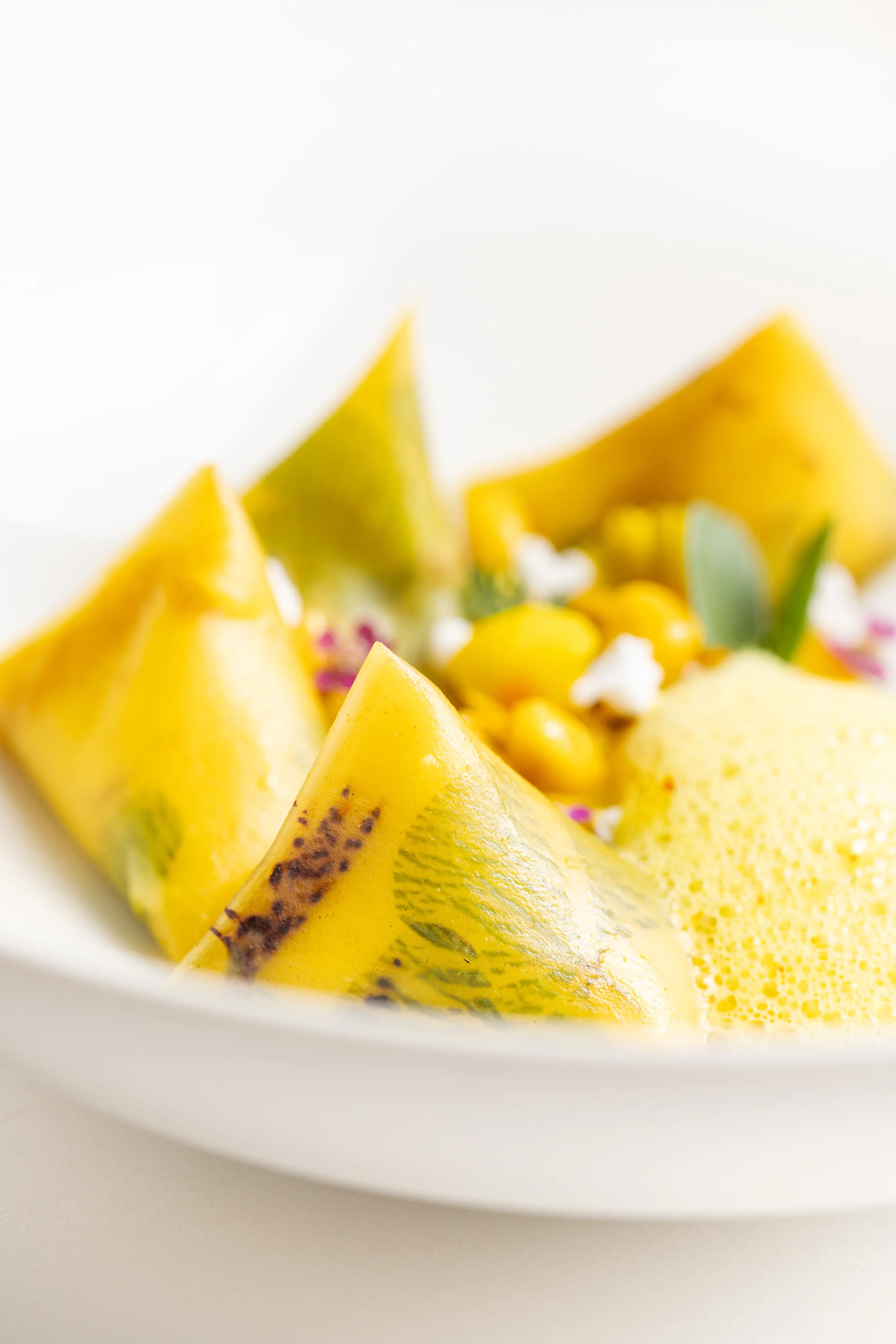 Assiette avec des raviolis jaunes décorés de feuilles, accompagnés de sauce mousseuse et garnis de petits légumes jaunes.