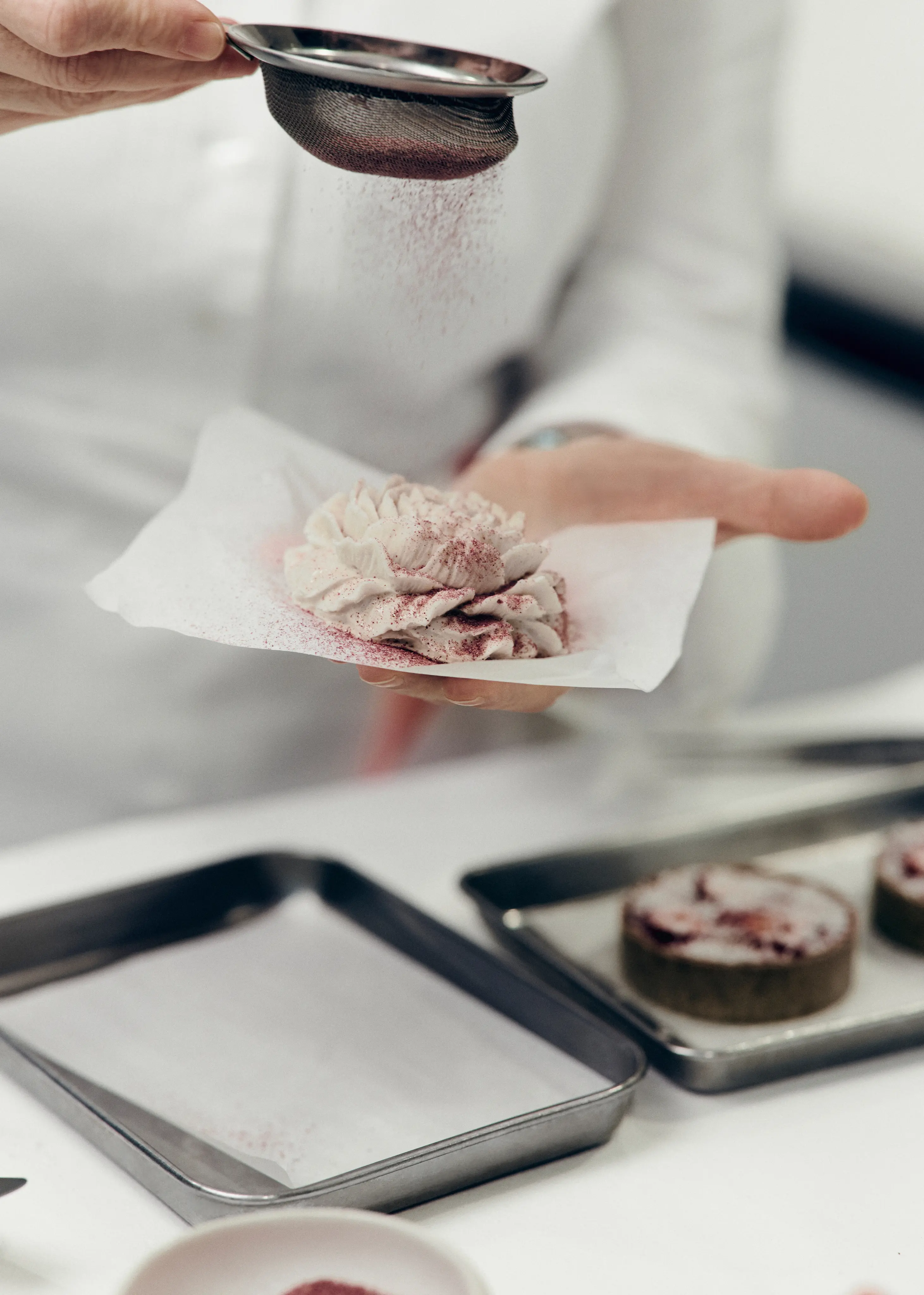 Main tenant un morceau de pâtisserie sur du papier, une autre main saupoudre de la poudre rose avec une passoire fine.