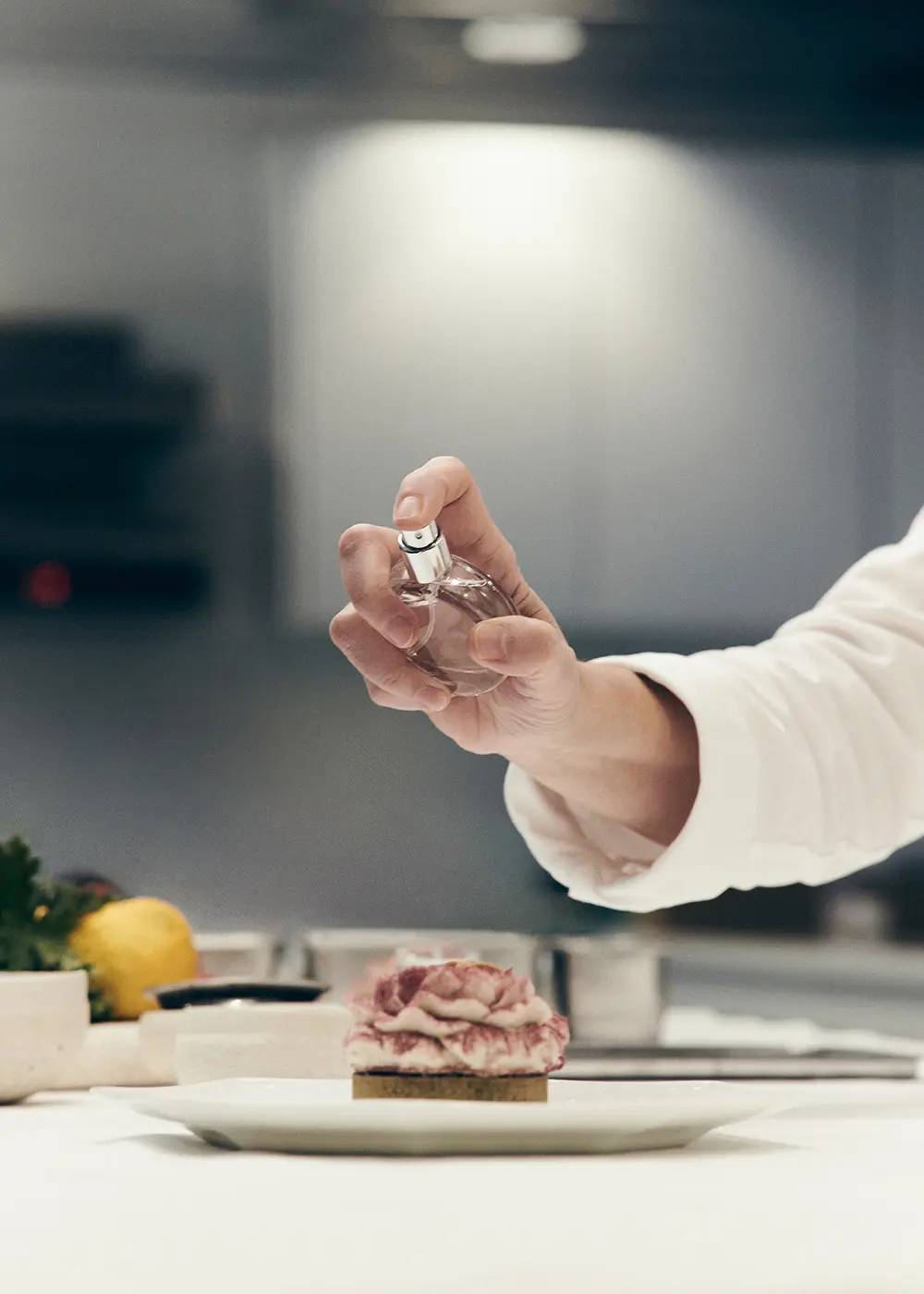 Une main tenant un flacon de parfum au-dessus d'une pâtisserie décorée sur une assiette blanche.