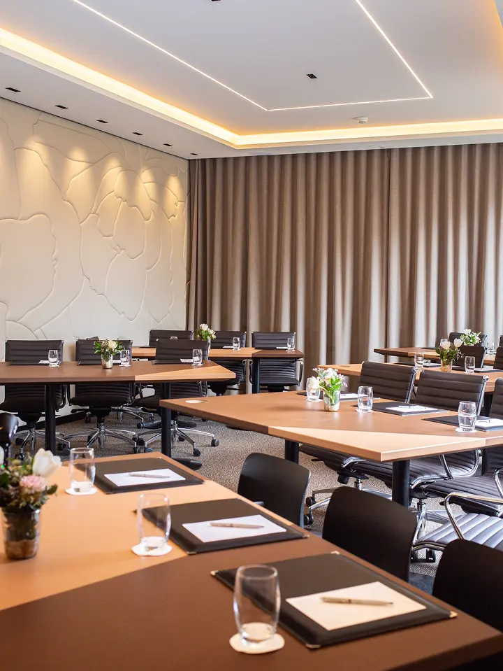 Salle de réunion moderne avec tables en bois, chaises noires, blocs-notes, stylos, verres d'eau et bouquets de fleurs.