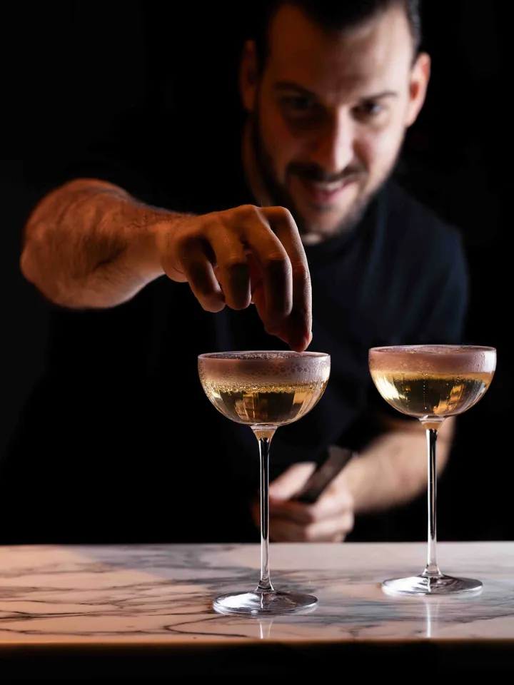 Un barman souriant garnit un cocktail doré dans un verre à pied sur un comptoir en marbre.