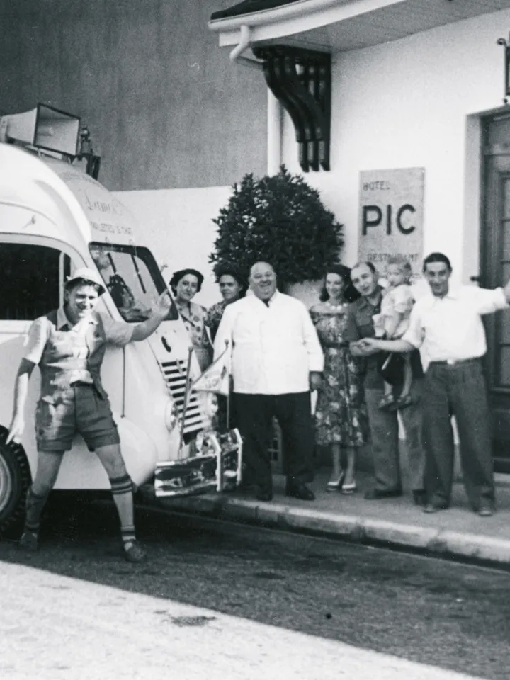 Photo en noir et blanc d'un groupe de sept personnes souriantes, dont un enfant, posant devant un véhicule et un bâtiment avec un panneau indiquant 'Hôtel Pic'.