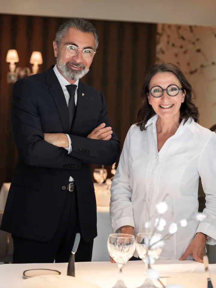 Un homme en costume noir avec une cravate et des lunettes, debout avec les bras croisés à côté d'une femme souriante en chemise blanche et lunettes, dans un cadre de restaurant élégant avec une table dressée devant eux.