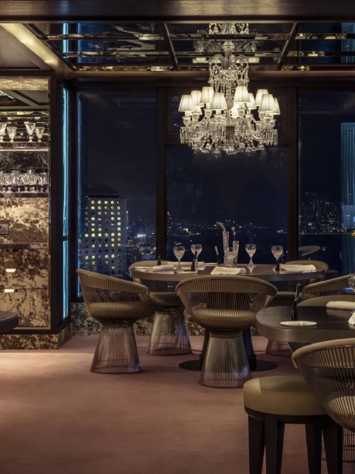 Salle à manger élégante avec grandes fenêtres donnant sur une ville de nuit, chaises en métal design et lustre en cristal.