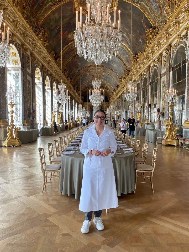 Personne en robe blanche et lunettes debout devant une longue table dressée dans une salle somptueusement décorée avec de grands lustres en cristal et des miroirs dorés.