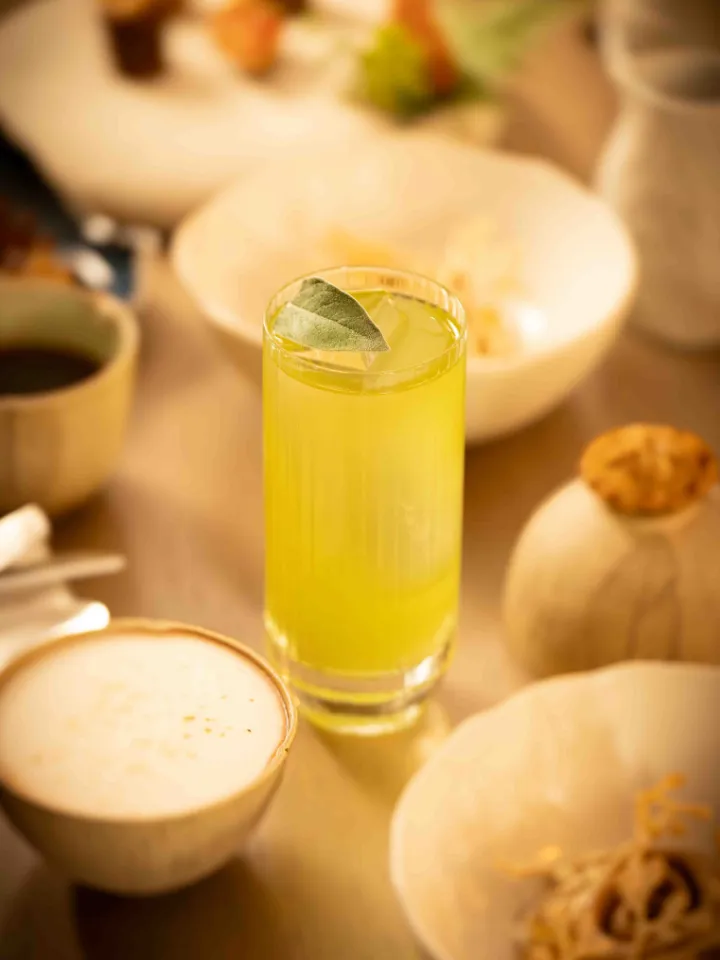 Verre de boisson jaune garnie d'une feuille verte sur une table avec des bols flous en arrière-plan.