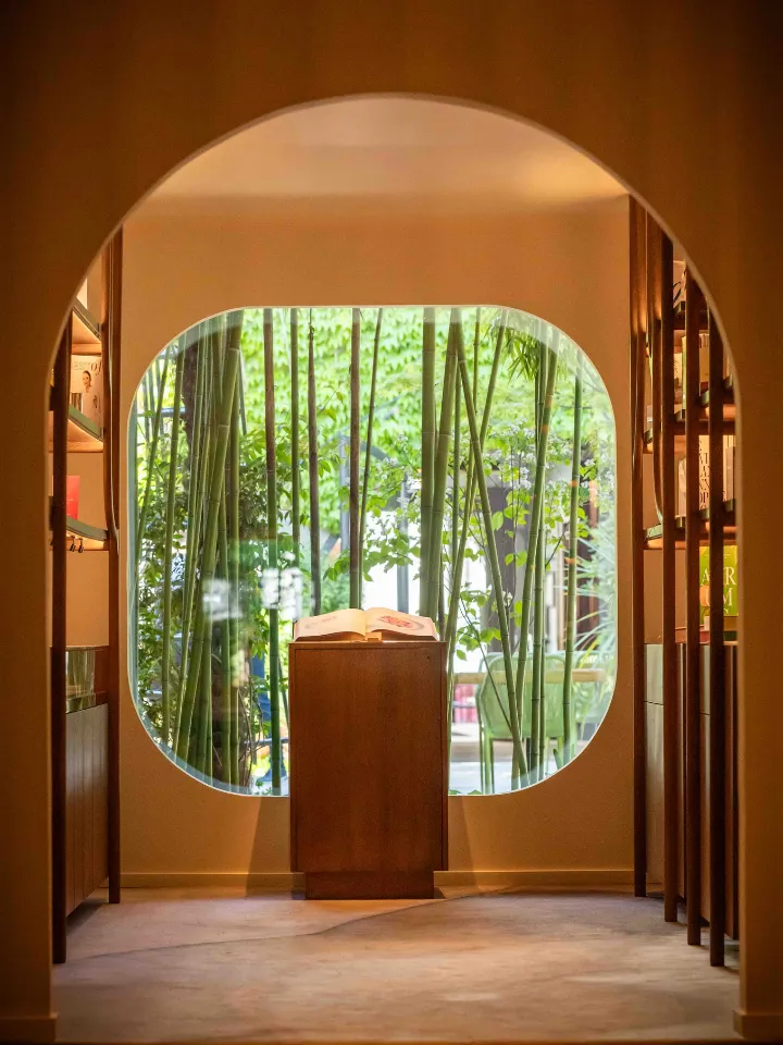 Innenansicht eines Raumes mit einem großen, abgerundeten Fenster mit grünem Bambus und einem Holzsockel mit einem offenen Buch.