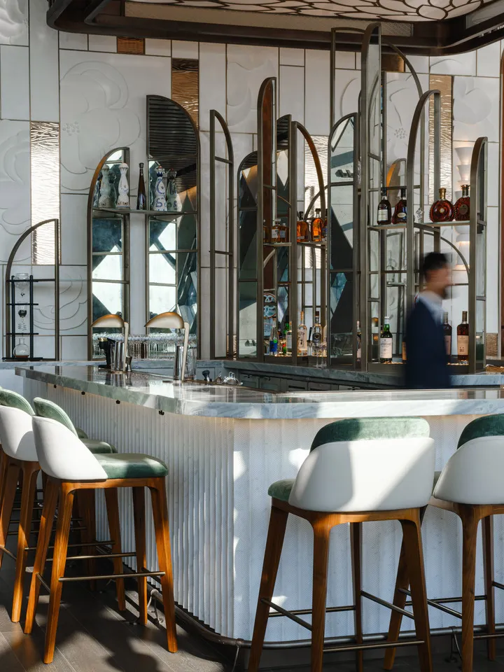 Bar élégant avec un comptoir en marbre, tabourets en bois et cuir vert, devant des étagères de bouteilles et miroirs décoratifs; un homme marche au loin.