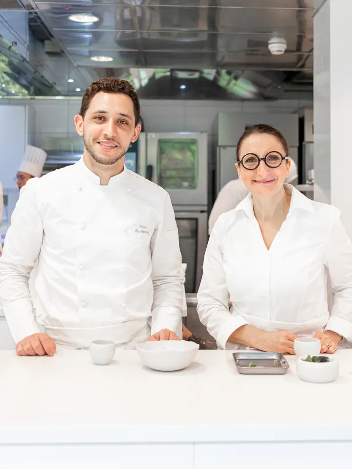 Deux chefs souriants, un homme en veste blanche de chef et une femme en blouse blanche avec lunettes rondes, posant dans une cuisine professionnelle.