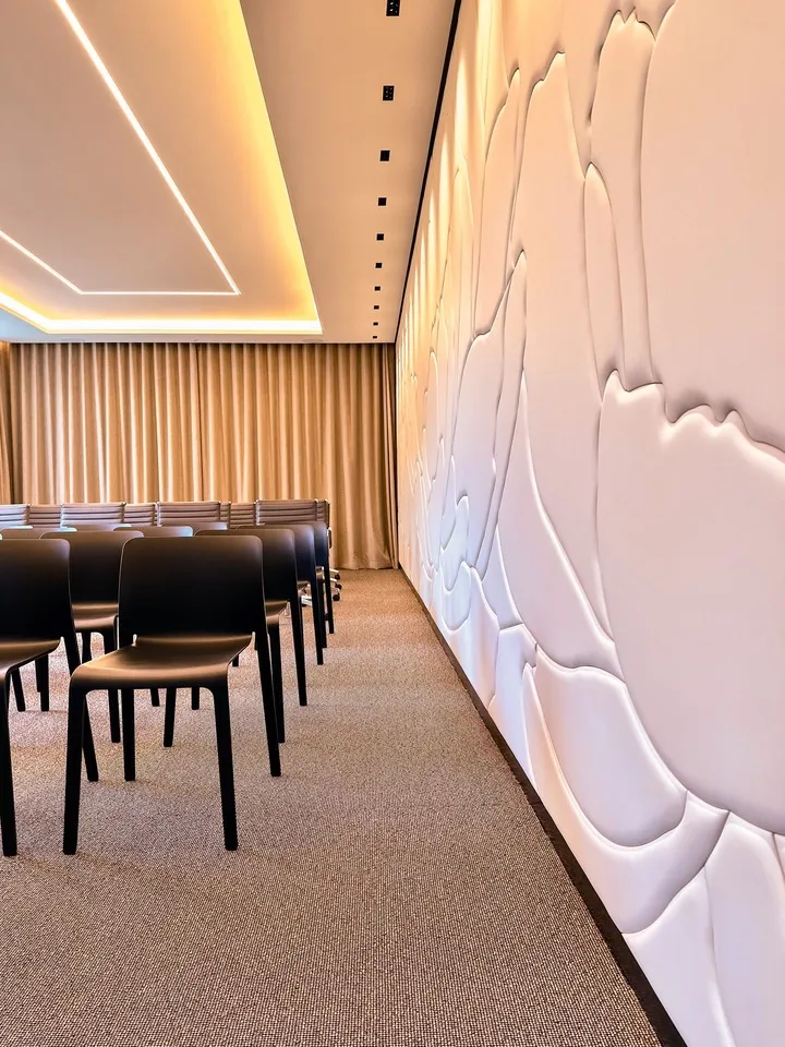 Intérieur d'une salle de conférence moderne avec rangées de chaises noires, mur texturé blanc et éclairage indirect au plafond.