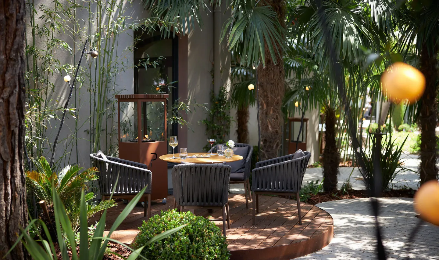 Coin repas extérieur avec une table ronde en bois entourée de quatre chaises grises sur une terrasse en bois, entouré de plantes vertes et de palmiers.