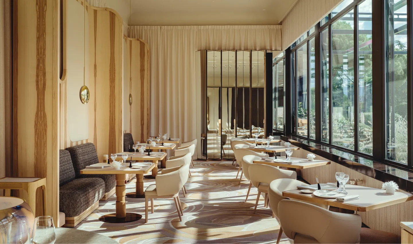 Salle de restaurant élégante avec tables en bois, chaises crème et grandes fenêtres donnant sur un jardin.