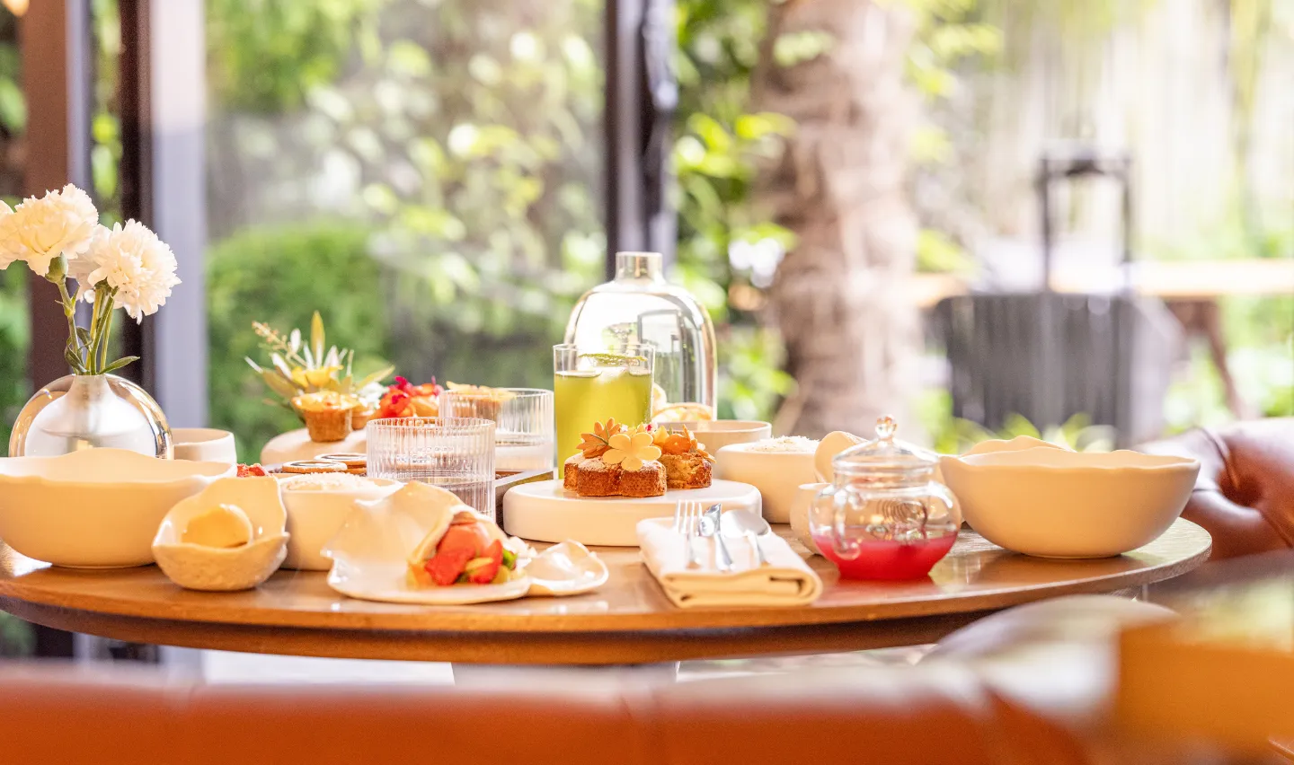 Table ronde en bois avec assortiment de pâtisseries, boissons et vaisselle blanche dans un cadre lumineux avec vue sur un jardin flou.