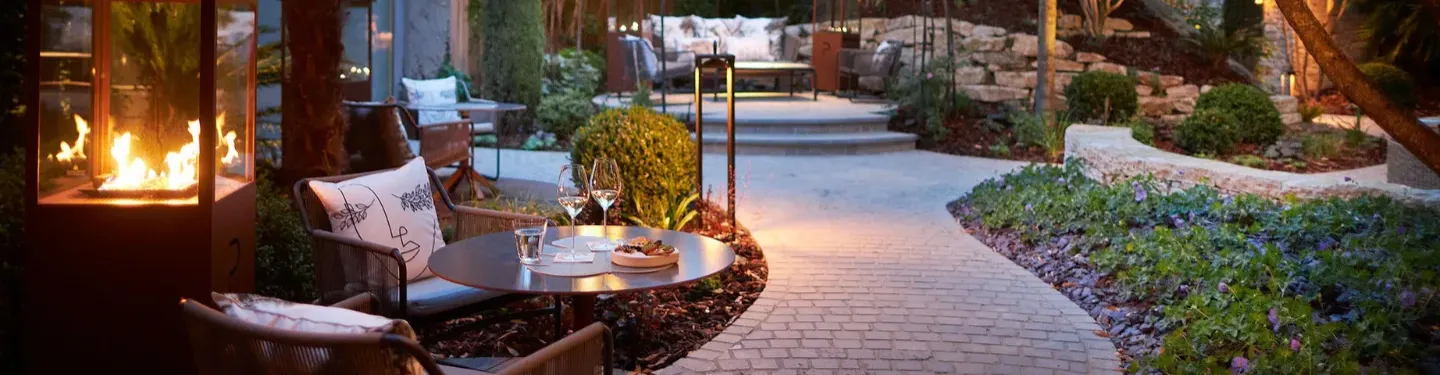 Terrasse extérieure cosy avec chemin pavé, table ronde dressée de verres et apéritifs, fauteuils à coussins et cheminée allumée au crépuscule.
