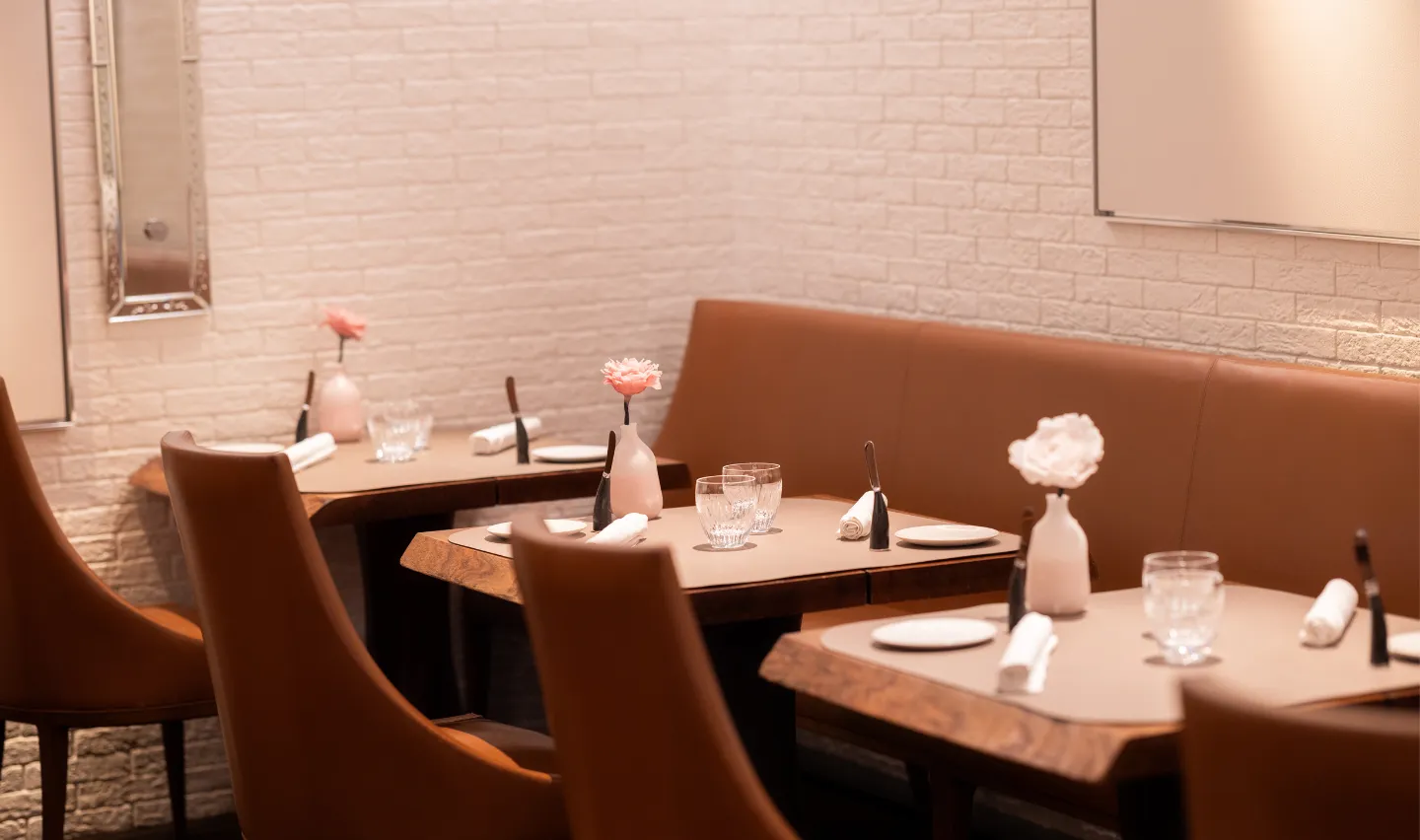 Tables dressées avec nappes, verres, serviettes roulées, assiettes et vases fleuris, dans un restaurant aux murs en briques blanches.