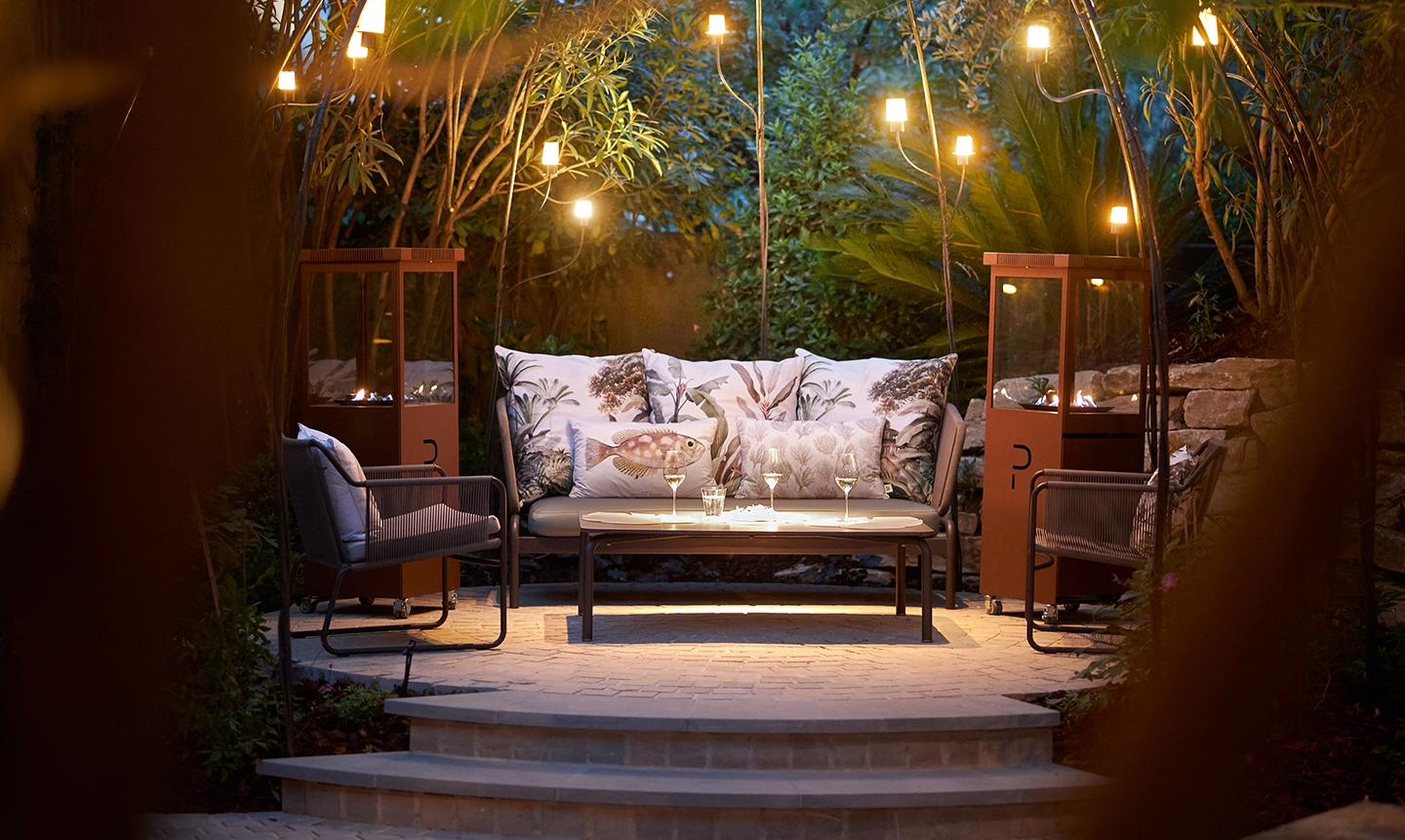 Beleuchteter Sitzbereich im Freien mit Sofa, Sesseln, dekorativen Kissen und Hängelampen, umgeben von viel Grün.
