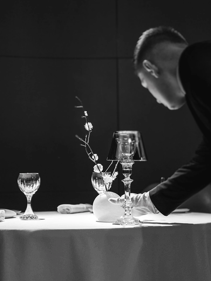 Demi-chef de rang en gants blancs dressant une table élégante avec verres en cristal et nappe blanche.